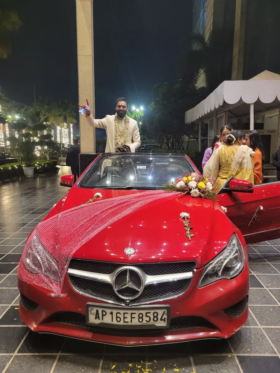 Groom beside a flower-decorated red wedding car at a nighttime celebration.