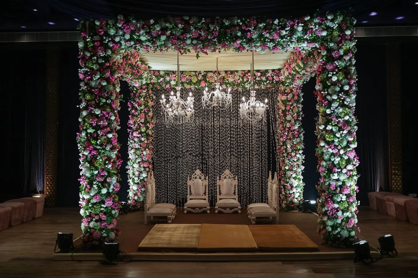 Floral wedding mandap framed by flower-covered arches and hanging lights.